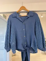 Camisa azul de manga larga con diseño abullonado, detalles de bordado estilo broderie en las mangas y laterales, cuello camisero, cierre frontal con botones y lazo para atar en el frente.