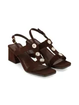 Sandalias de taco cuadrado en tono marrón chocolate, confeccionadas en gamuza. Poseen tiras cruzadas en el empeine con detalles de botones metálicos dorados decorativos, tira ajustable al talón con hebilla dorada y taco ancho estable.