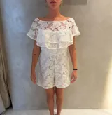 Mono corto de encaje blanco con escote off-shoulder y volados superpuestos en el pecho.
