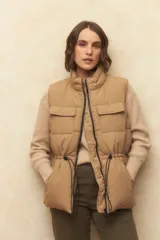 Chaleco puffer color crema, con cuello alto, cierre frontal y cuatro bolsillos con solapa. Cuenta con cintura ajustable con cordón.