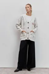 Cardigan de punto color crema con cuello en V y cierre frontal de botones. Presenta bordados coloridos con motivos variados como flores, cerezas, estrellas y frases en toda la prenda. Incluye dos bolsillos frontales.