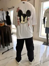Remera oversize blanca de manga corta, con cuello redondo y etiqueta decorativa en la parte posterior del cuello. Presenta un gran estampado en la espalda que combina un diseño estilizado de Mickey Mouse con caracteres en estilo asiático en color rojo.