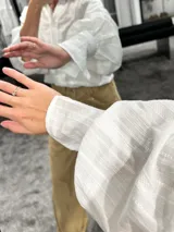 Camisa blanca de textura rayada con cuello mao y cierre frontal de botones. Presenta mangas largas abullonadas con puños anchos y corte holgado.