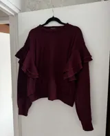 Sweater de punto color bordó con diseño de volados en los hombros y mangas largas. Presenta una textura acanalada fina y corte holgado.