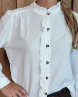 Camisa blanca de algodón con cuello alto tipo mao y volados sutiles a lo largo de la botonadura frontal. Presenta mangas largas con frunces en los hombros y botones frontales.