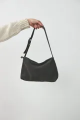 Cartera de hombro mediana confeccionada en cuero agamuzado color gris, con cierre superior y correa ajustable mediante hebilla metálica.