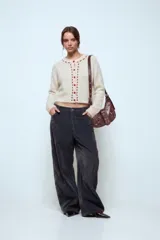 Cardigan de punto color beige, de corte cropped y manga larga. Presenta cierre frontal con botones y detalles de pespuntes decorativos en color bordó a lo largo de la abertura y el cuello.