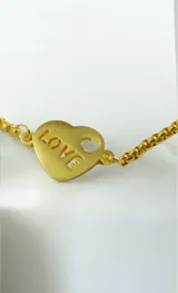 Pulsera de cadena tipo veneciana en tono dorado con sistema de cierre ajustable mediante deslizador. Presenta un dije en forma de corazón con la palabra LOVE grabada y un pequeño cristal incrustado.
