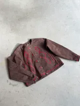 Chaqueta de gamuza color chocolate con detalles de bordado en color rojo, cuello redondo, cierre frontal con botones y mangas largas con puños abotonados.