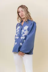 Camisa azul de calce relajado con bordado floral blanco en el frente. Presenta mangas largas amplias con ligero fruncido en el hombro y cierre frontal con botones.