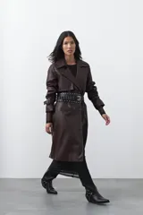 Trench coat de símil cuero color marrón oscuro, con solapas amplias, cierre frontal con botones, cinturón ajustable en la cintura y mangas con tiras ajustables. Presenta un largo por debajo de la rodilla y bolsillos laterales.