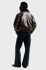 Campera de estilo aviador confeccionada en tejido revestido con acabado de cuero sintético. Presenta cuello de sherpa en contraste, cierre frontal de cremallera, hombros caídos y bolsillos delanteros de parche con solapa y botón a presión. Incluye elástico fruncido en el bajo y puños ajustables con botón a presión.