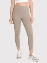 Calza deportiva de tiro alto con largo al tobillo, confeccionada en tejido elástico de color beige. Presenta un diseño minimalista sin bolsillos y un pequeño logo bordado en la cadera.