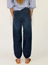 Pantalon de denim azul oscuro con diseño abombado y puños fruncidos con elástico en los tobillos. Cuenta con bolsillos laterales y cierre frontal con botón y cremallera.