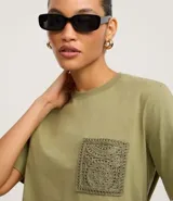 Remera de manga corta y cuello redondo color verde oliva, confeccionada en algodón, con un bolsillo cuadrado aplicado en el pecho con diseño de crochet intrincado.