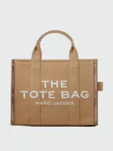 Bolso tipo tote bag de Marc Jacobs en color beige, confeccionado en lona resistente. Presenta el texto "THE TOTE BAG" y "MARC JACOBS" impreso en la parte frontal en color negro. Cuenta con asas superiores y una correa de hombro ajustable y extraíble, además de ribetes laterales negros con inscripciones.