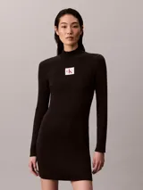 Vestido mini de punto acanalado color negro, de manga larga y cuello alto. Cuenta con una etiqueta con el logo de Calvin Klein en el centro del pecho.