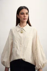Camisa a rayas verticales color beige y blanco, con cuello clásico, cierre frontal con botón dorado, mangas largas abullonadas con puños elásticos, dos bolsillos frontales y corte holgado.