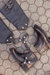 Bota de caña alta confeccionada en cuero color marrón caoba con acabado desgastado. Presenta una suela track robusta, correas con hebillas en la parte superior de la caña y un arnés decorativo con herrajes metálicos en el tobillo.