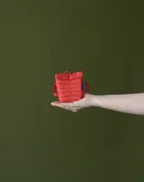 Charm en forma de mini mochila color rojo, confeccionada en material acolchado con costuras horizontales y tira lateral con etiqueta de la marca. Cuenta con una pequeña argolla metálica en la parte superior para colgar.
