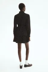Vestido corto negro con estampado de lunares blancos, escote en V con volados, mangas largas abullonadas y cordón ajustable en la cintura.
