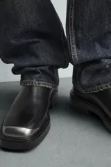 Bota tipo Chelsea de cuero negro con paneles elásticos laterales, suela de goma con volumen y detalle de pieza metálica en la puntera.