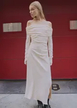 Vestido largo de color crema con diseño drapeado, escote palabra de honor y mangas largas con puños drapeados. Presenta una abertura lateral en el bajo y cierre de cremallera lateral.
