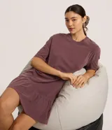 Vestido tipo camiseta de algodón, color malva o ciruela con efecto lavado, de corte recto y holgado, con cuello redondo y mangas cortas. Presenta un volado fruncido en el bajo.