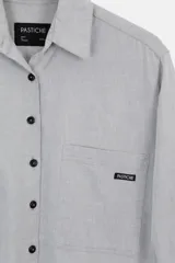 Camisa oversized de algodón color gris claro, con cuello clásico, botones negros y bolsillo frontal con etiqueta de marca.