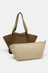 Cartera shopper de hombro confeccionada en gamuja color marrón, con diseño estructurado de laterales abiertos y doble asa superior. Cuenta con cierre de botón imantado e incluye un bolsillo interior extraíble con cremallera.