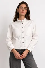 Campera de algodón color blanco, con cuello alto con volados, cierre frontal de botones plateados, mangas largas y cuatro bolsillos frontales.