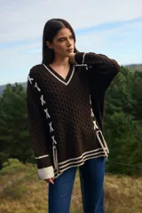 Sweater de punto estilo preppy en color crudo con detalles en marrón. Presenta cuello en V con ribete a rayas, tejido con textura de ochos y cordones laterales decorativos. Posee puños y ruedo acanalados con diseño a rayas.