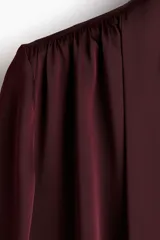 Blusa de manga larga color borgoña, confeccionada en tela satinada o gasa con caída. Presenta cuello alto con lazo o moña larga para atar, y frunces en los hombros. Las mangas terminan en puños ajustados.