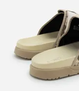Ojota tipo slide con estampa camuflada en tonos beige y marrón. Presenta dos tiras ajustables con cierre de velcro y suela de goma antideslizante.