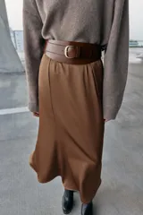 Cinturón tipo fajín de cuero con diseño asimétrico y ondulado. Presenta un acabado liso en tono marrón oscuro y cierre mediante piezas a presión.