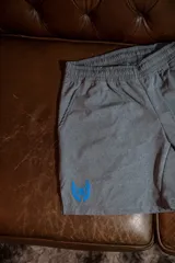 Short deportivo gris melange con cintura elástica y aberturas laterales para mayor movilidad. Presenta un logo de alas estampado en color azul en la parte inferior izquierda.