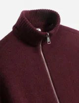 Poncho tejido color bordó con cuello alto y cierre frontal completo. Presenta mangas cortas con terminación en puño acanalado y un diseño holgado.