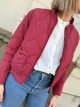 Campera tipo bomber acolchada con diseño de pespuntes ondulados, cierre frontal y bolsillos laterales con botón. Presenta un corte moderno y acabado en color bordo.
