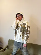 Hoodie oversize de color blanco con estampado artístico en tonos marrones y dorados en el pecho y las mangas. Presenta un forro interior de capucha en tejido marrón texturizado y costuras reforzadas.
