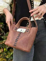 Cartera de cuero marrón de formato estructurado, con doble asa de mano y correa larga ajustable para llevar al hombro o cruzada. Cuenta con detalles de costuras negras a la vista y una pieza decorativa central de resina con efecto carey y aplique metálico.