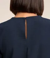 Blusa blanca de poliéster con cuello redondo, mangas cortas, cierre trasero en forma de gota, pequeñas aberturas laterales y acabados en pespuntes en las mangas y el cuello.