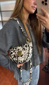 Bandolera con silueta de media luna y diseño estructurado. Confeccionada en cuero con estampado animal print tipo vaca, cuenta con cierre superior, herrajes metálicos y placa frontal dorada con logo. Incluye correa regulable para llevar al hombro o cruzada.