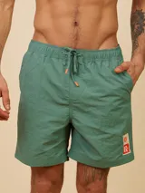 Short de baño color verde, con cintura elástica y cordón ajustable. Presenta un parche decorativo con logo en la parte inferior de la pierna izquierda.