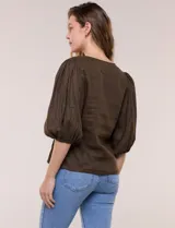 Blusa color beige confeccionada en lino, con escote en V pronunciado y cierre frontal de múltiples lazos para atar. Presenta mangas abullonadas tres cuartos con puños elásticos y corte relajado.