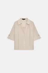 Camisa beige de tejido texturizado, con cuello solapa, escote en V y cierre frontal con botones. Presenta mangas cortas con vuelta y trabilla decorativa.