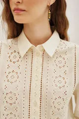 Camisa de algodón color crudo con diseño de encaje calado y detalles de plisados verticales en el frente. Presenta cuello clásico, cierre frontal de botones y mangas largas con puños delicados.