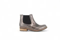 Botín tipo Chelsea de cuero metalizado en tono bronce, con paneles elásticos laterales en color beige. Presenta detalles de costuras decorativas tipo brogue en la puntera y laterales, con suela de goma antideslizante y taco bajo foliado.