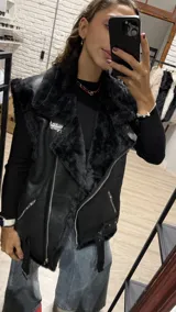 Chaleco estilo gamulán de cuero sintético negro con interior y bordes de corderito sintético color crema. Presenta cierre frontal, bolsillos laterales con cierre y cinturón ajustable en el ruedo.