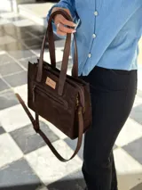 Cartera tipo tote de gamuza color chocolate, con doble asa de mano y correa larga ajustable y extraíble. Presenta un bolsillo frontal con cierre metálico y una etiqueta de cuero con el logo de la marca en el frente.