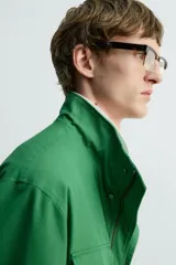 Chaqueta tipo cazadora de corte relajado en color verde, confeccionada en tejido de mezcla de algodón. Presenta cuello subido con forro interior a cuadros, manga larga con puños abotonados y cuatro bolsillos frontales de plastrón con solapa. Incluye cierre frontal de cremallera oculta por solapa con botones a presión y cintura ajustable en el interior.